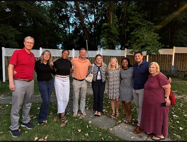 April smith with a group of nine diverse adults stand side-by-side in a backyard, smiling and posing for a photo at dusk. They are arranged in a horizontal line in front of a white lattice fence with dark green trees in the background.