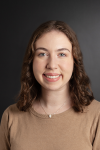 A white woman with shoulder length curly brown hair and blue eyes smiles at the camera. She is wearing a light brown shirt and is in front of a gray background.
