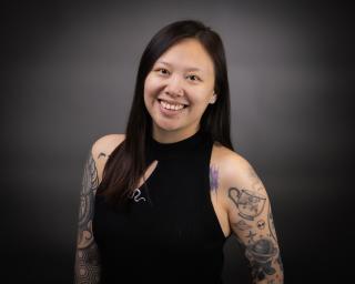 Asian woman with short brown hair and a lot of arm tattoos wearing a black shirt