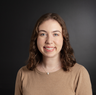 A white woman with shoulder length curly brown hair and blue eyes smiles at the camera. She is wearing a light brown shirt and is in front of a gray background.